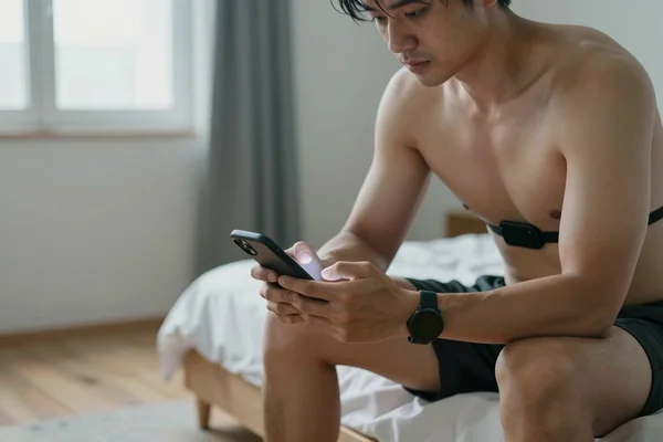 Man sitting on the edge of his bed in the morning with a chest strap and sports watch, checking his HRV on his phone