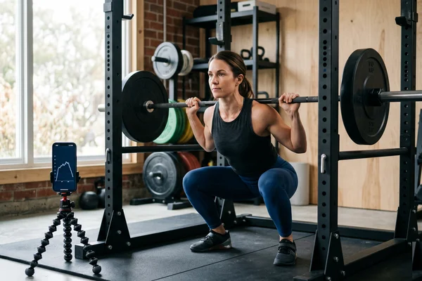 Athlete doing a controlled barbell squat with two reps in reserve, a velocity-tracking app on a phone beside the rack — setting load without a max test.